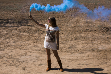 young beautiful woman holding a blue smoke bomb outdoors. Brown background. Sunny. Casual clothing. Lifestyle