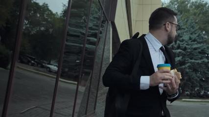 Young angry busy man with an unhealthy lifestyle eating a sandwich, drinking coffee and talking on mobile phone while walking in a hurry on the street