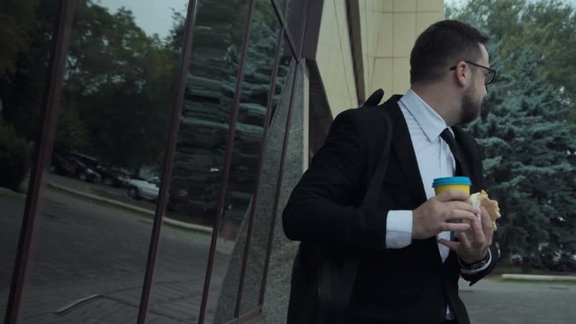 Young angry busy man with an unhealthy lifestyle eating a sandwich, drinking coffee and talking on mobile phone while walking in a hurry on the street
