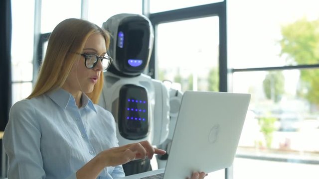 Cheerful girl holding laptop and talking to robotic machine