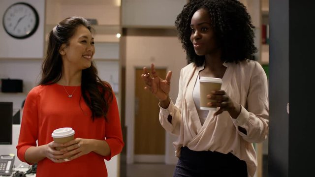 Businesswomen talking & holding cups of coffee walking through busy office