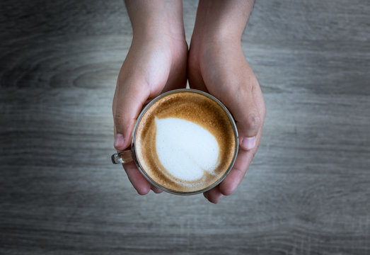 Female hands holding a cup of coffee with heart make up on face of milk cream on wooden table, top view