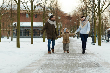 Little girl holding by her parents hands while taking walk in winter park