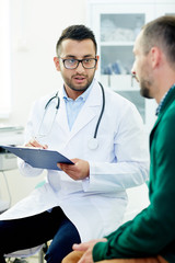Young clinician making notes in medical card of patient while talking to him