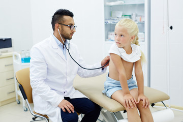 Little girl sitting on couchette while physician with stethoscope examining her