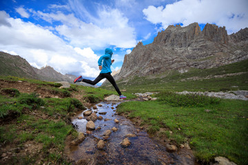 young woman trail runner jumping over stream water at mountain
