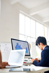 Busy manager texting in smartphone while sitting in front of computer monitor with financial chart and graph