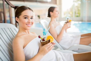 Smiling girl wrapped into white towel enjoying exotic cocktail from coconut while relaxing on deckchair