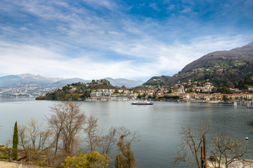 Fototapeta na wymiar Lake Maggiore, Laveno, Italy. Picturesque view of the lake promenade that leads to the harbor, the ferry boat and the church. In the background the Alps with some snowy peaks, on a pleasant winter day