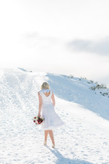 Fototapeta na wymiar Blonde Frau in weißem Kleid mit Brautstrauß geht in Winterlandschaft