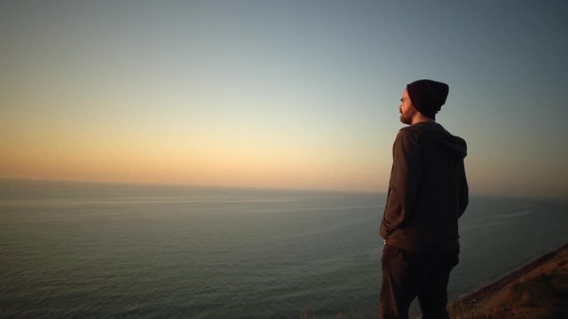 picture of handsome bearded guy in hat standing on the top of hill over sea looking ahead have feeling of freedom during magnificent sunrise side view slow motion