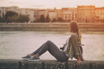 Young woman with camera and backpack enjoys sitting at the riverbank and looking at the city. 