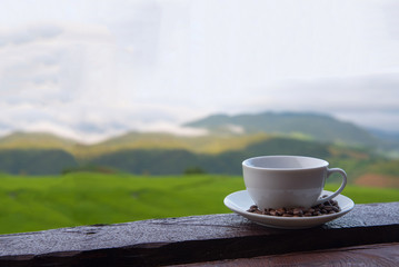 A white cup of hot coffee and coffee beans put interspersed in dish on wood table with green rice field background,wallpaper
