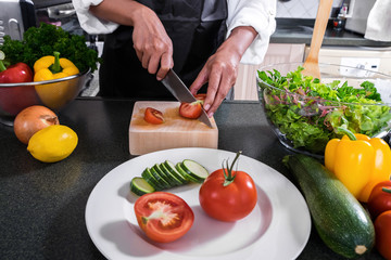 Close up of woman hand cutting tomato on chopping wood board with sharp knife and cooking vegetables salad in kitchen