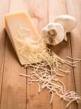 Garlic and Parmesan Cheese Wedge with Shredded. an angled view of a wedge of parmesan cheese and two cloves of garlic on wood with shredded cheese