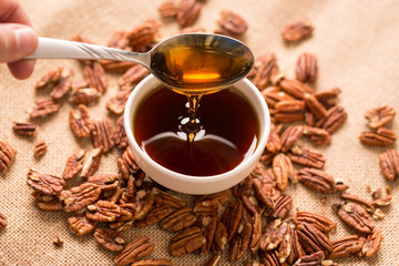 Dipping Spoon in Maple Syrup with Pecans. a close up view of a bowl of maple syrup with a spoon dipped in and pouring some out surrounded by pecans on a textured brown cloth