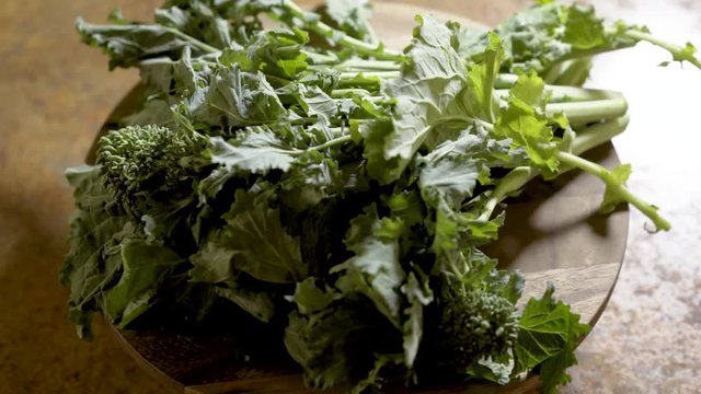 Clusters of stalks of fresh rapini on a round cutting board rotating in beautiful backlit light.