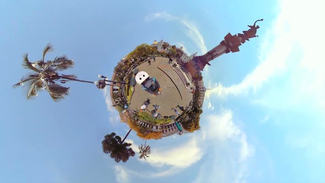 A spherical 360 tiny-planet view of the Plaza Grande in Quito, Ecuador, the Independence monument, colonial buildings, palm trees and people walking in a rotating mini-planet effect under a blue sky.