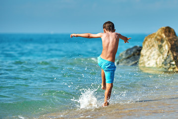 Cute Caucasian boy is running in the water along the sea shore. Back view.