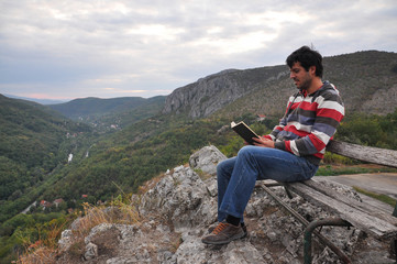Naklejka na ściany i meble Man sitting and reading on the edge of a cliff surrounded by nature. Concept of peace in nature