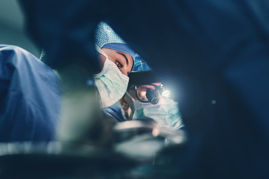 Surgeon doctor operating using special lamp lighting and glasses loupes wearing blue surgical mask and surgical cap in surgery room with his team surgeons operating live heart beating heart surgery