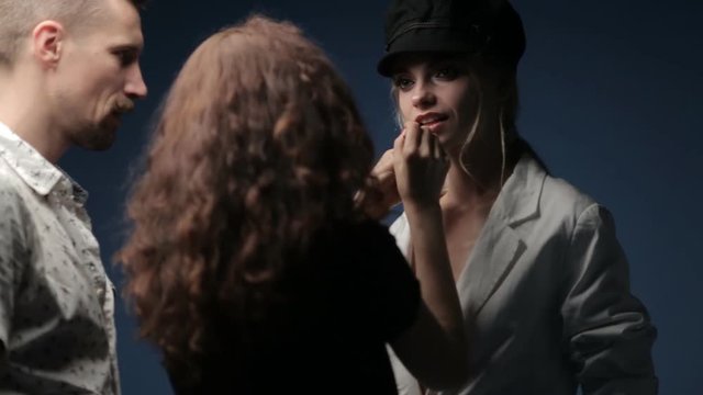 A make-up artist and a stylist work with a model in a black hat