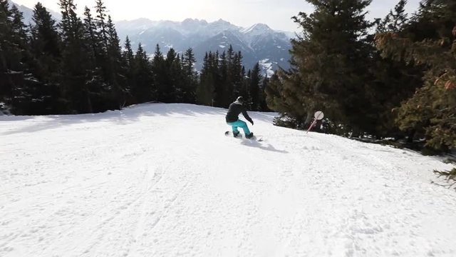 Slow motion of a snowboarder snowboarding downhill on the ski slope with his snowboard and turning with beautiful view of mountains and sunshine