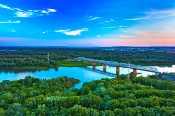 A railway bridge across a calm river surrounded by green magical forest at sunset with a beautiful...