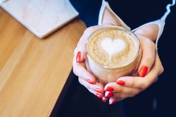 Fototapeta na wymiar A girl with red nails is holding a cup with cappuccino with a pattern in the form of a heart