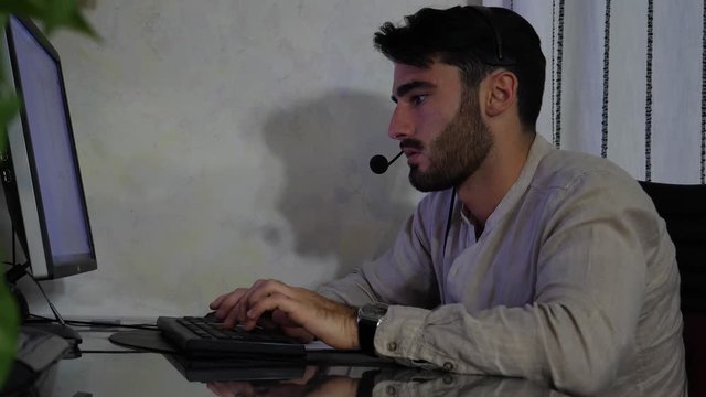 Young male home worker talking with customer or client through headset and microphone, working from his living room in front of computer late at night