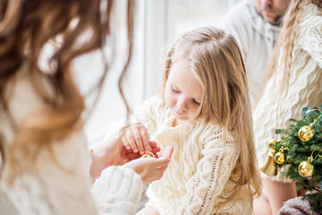 A blonde mother and daughter in white knitted sweaters decorate a small Christmas tree with golden toys. A young woman shows in hands the girl New Year decorations. Copy space. Festive atmosphere card