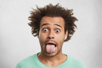 Young funny Afro American man looks surprisely, makes grimace, shows tongue as quarrels with wife....