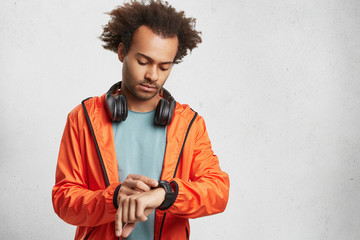 Portrait of dark skinned male student wears orange anorak, looks at watch time, waits for friends to go together on football match, isolated over white background. Hipster guy with headphones © WHstudio Leushin N
