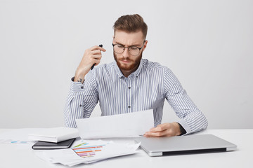 Horizontal portrait of attractive beatded male manager, sits at office, surrounded with laptop, documents, reads attentively financial report, makes corrections, going to present on business meeting