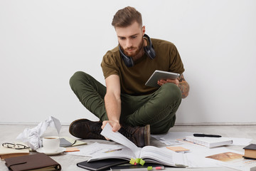 Preoccupied beared male college student with trendy hairdo looks attentively into book, holds...