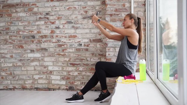 Resting woman takes selfie after hard work out. She sits by the window and takes photos on the smartphone.