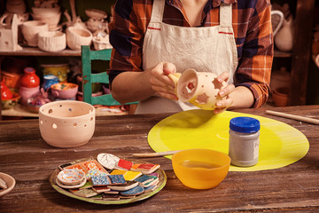 Close-up of a woman potter in a plaid shirt and apron paints a wooden tassel in a gray clay candlestick with asterisks on a wooden table on which there are tools, clay samples, and in  in the workshop