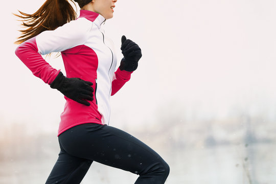 Winter running athlete woman on cold run jogging fast with speed and sprint on outside workout wearing warm clothing gloves, winter tights and wind jacket in snow weather.