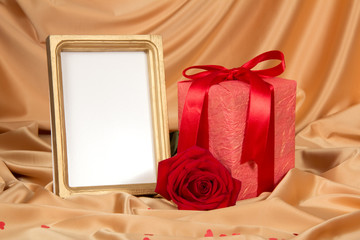 Obraz na płótnie Canvas Box with gift, scarlet rose and frame for photo on background of beige silk fabric