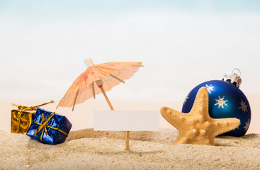 A blank card on peg, an umbrella, Christmas ball and gifts, starfish in the sand against the background of the sea.