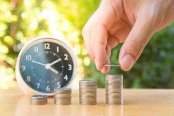Fotobehang Human hand is putting coin to growing coin stack with clock and nature background for saving money concept © magneticmcc