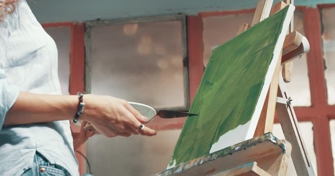 Attractive young female artist paints with acrylic on canvas in green tones in her studio