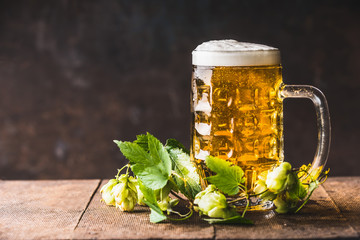 Beer mug with cap of  foam on table with fresh hops at dark rustic background, front view