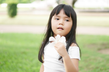 Asian children cute or kid girl holding white plumeria flower and make face fragrance on nature garden and green grass with tree for holiday relax on soft focus