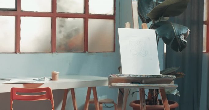 Pan video of an artist's studio with brushes, pencils, markers and other tools and a blank canvas on an easel