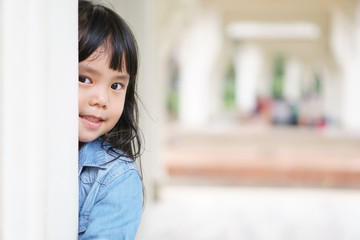 Asian children cute or kid girl wear jeans for fashion and playing hide and seek with sneaked or dodge behind the post and smile white teeth in the pavilion at park garden on relax holiday with space