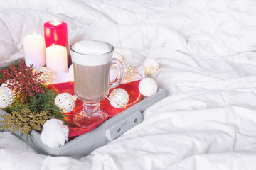 Breakfast with a cup of tea, a biscuit on a tray on the bed on a white blanket in the winter morning decorated with Christmas decorations, spruce branches with luminous garlands, the concept holiday