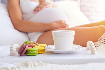 Macaroons with a cup of coffee on tray, pregnant woman sitting on back background