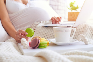 Hand of pregnant woman holding macaroon while using laptop in bed