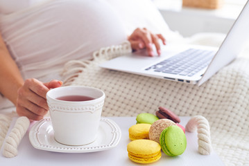Pregnant woman using laptop with tray of colorful macaroons and tea in bed